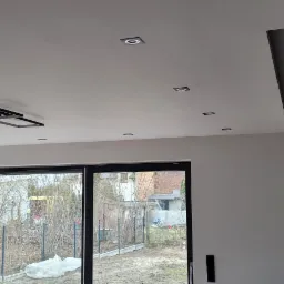 Nowoczesne oświetlenie sufitowe w nowo wykończonym salonie z dużymi oknami, widok na ogród z zewnątrz.