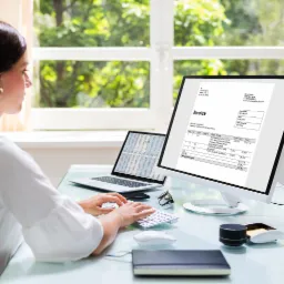 Kobieta pracuje przy biurku, na monitorze widoczna faktura, obok laptop z arkuszem kalkulacyjnym, na biurku notatnik, okulary i głośnik.