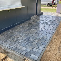 ProBrick Andrzej Majewski - Świeżo ułożona, wilgotna kostka brukowa w odcieniach szarości przed budynkiem z szarą elewacją, widoczny fragment chodnika i zaparkowany biały bus w tle.