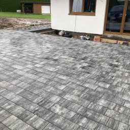 ProBrick Andrzej Majewski - Ułożona szara kostka brukowa na posesji, widoczny fragment domu z oknem i drzwiami tarasowymi, obok ściany leżą puszki farby i torba papierowa.
