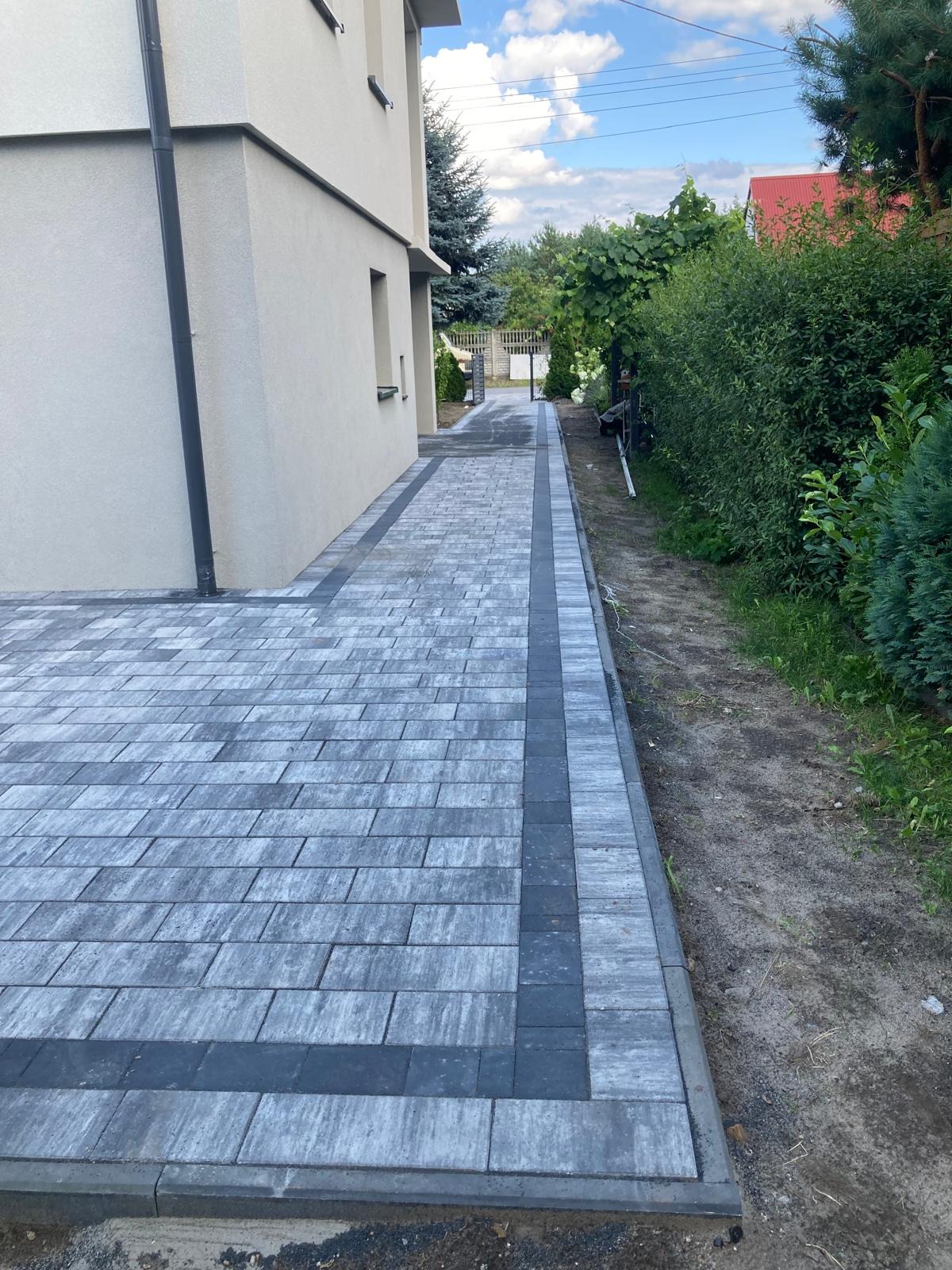 Ułożona szara kostka brukowa z ciemniejszym obramowaniem prowadząca wzdłuż jasnego budynku, widok na zielony ogród i błękitne niebo.
