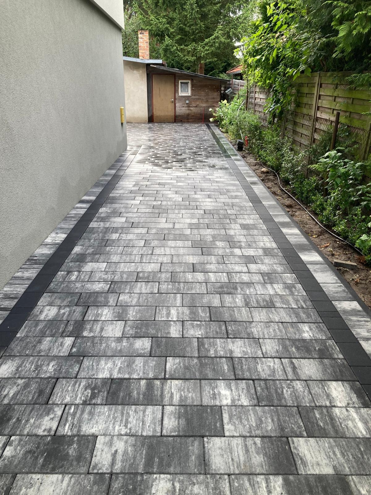 Ułożona kostka brukowa w odcieniach szarości z ciemnoszarą obwódką prowadząca do drewnianego budynku gospodarczego w Gorzowie Wielkopolskim. Widoczne ogrodzenie z siatki po prawej stronie...