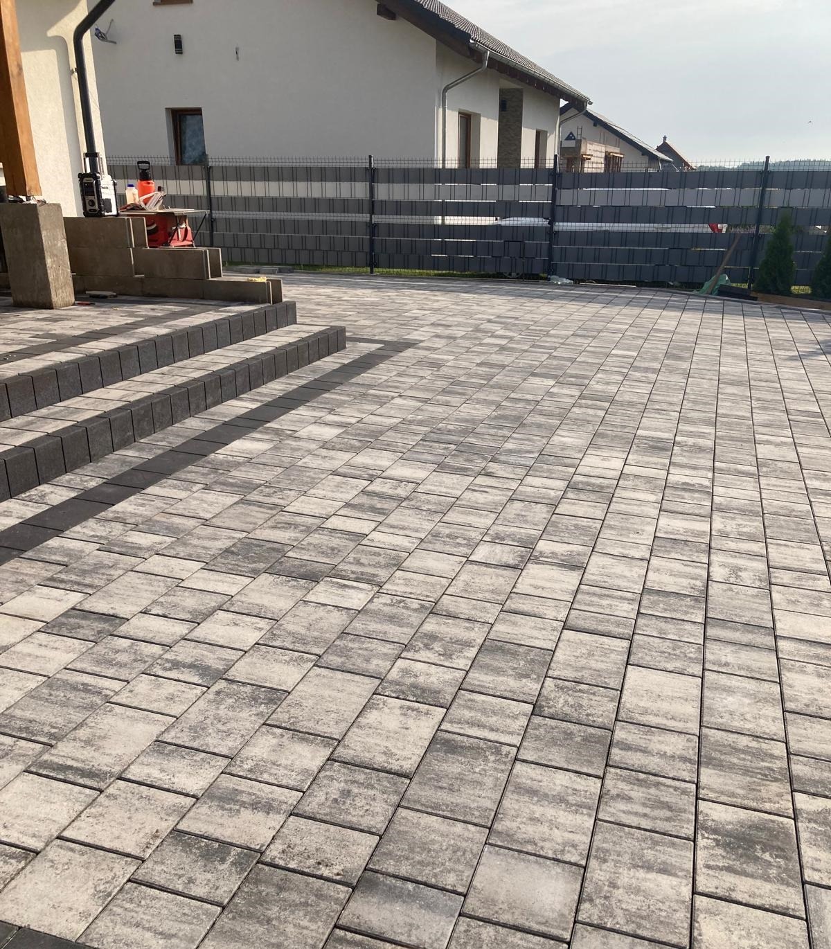 Nowoczesna nawierzchnia z kostki brukowej w odcieniach szarości, prowadząca do schodów i ogrodzenia panelowego z podmurówką, z widocznymi narzędziami na schodach.