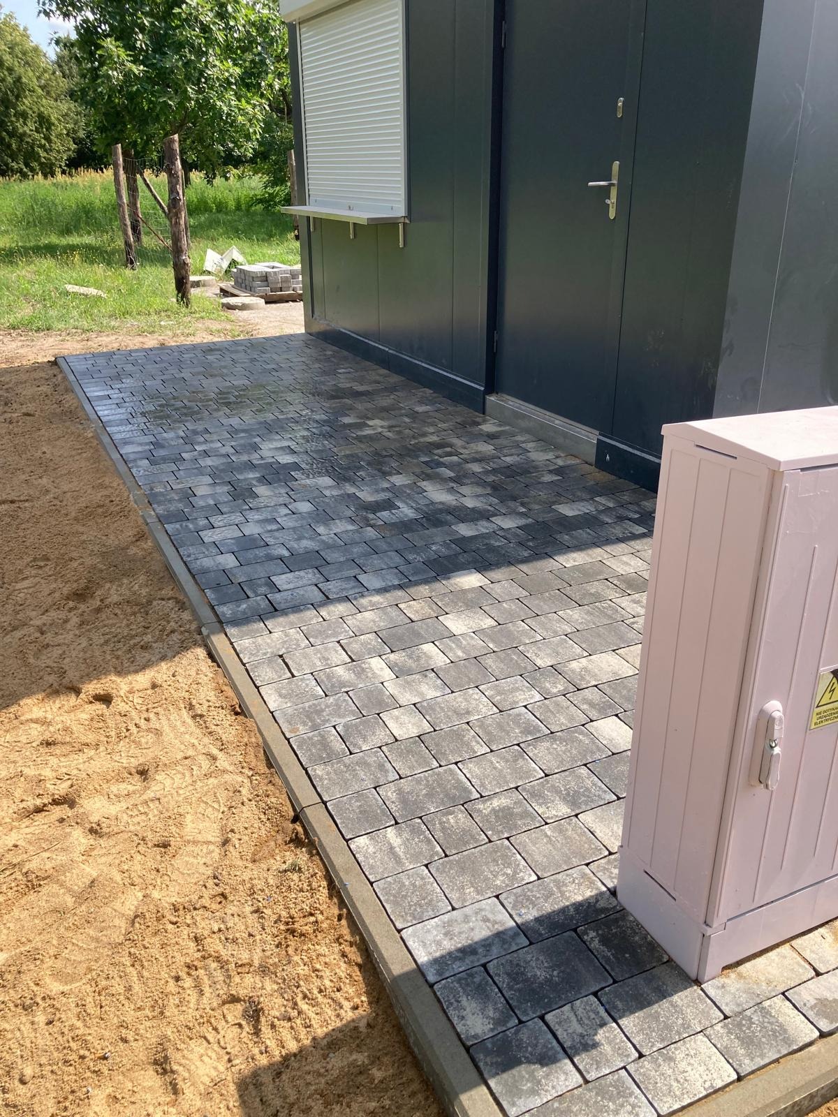 Ułożona szara kostka brukowa przed nowoczesnym budynkiem z metalową elewacją i roletą, widoczna skrzynka elektryczna w kolorze różowym.