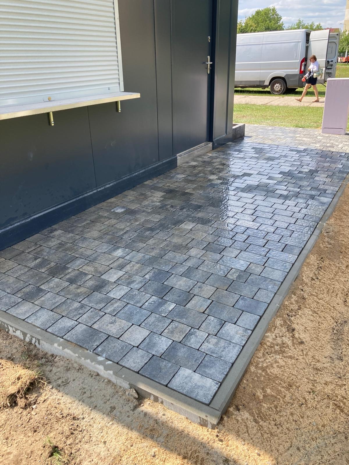 Świeżo ułożona, wilgotna kostka brukowa w odcieniach szarości przed budynkiem z szarą elewacją, widoczny fragment chodnika i zaparkowany biały bus w tle.