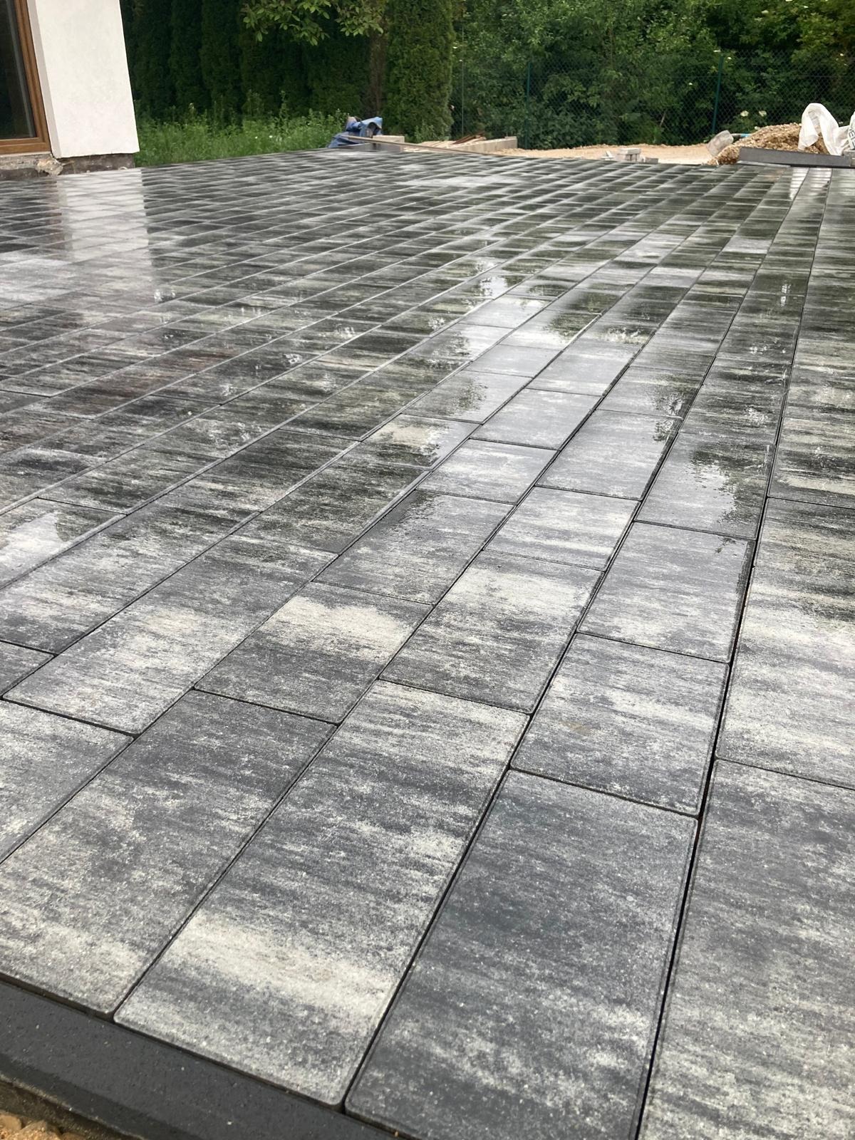Świeżo ułożona, mokra kostka brukowa o nieregularnym wzorze szarości i bieli, widoczne narzędzia w tle, tuż przy budynku.