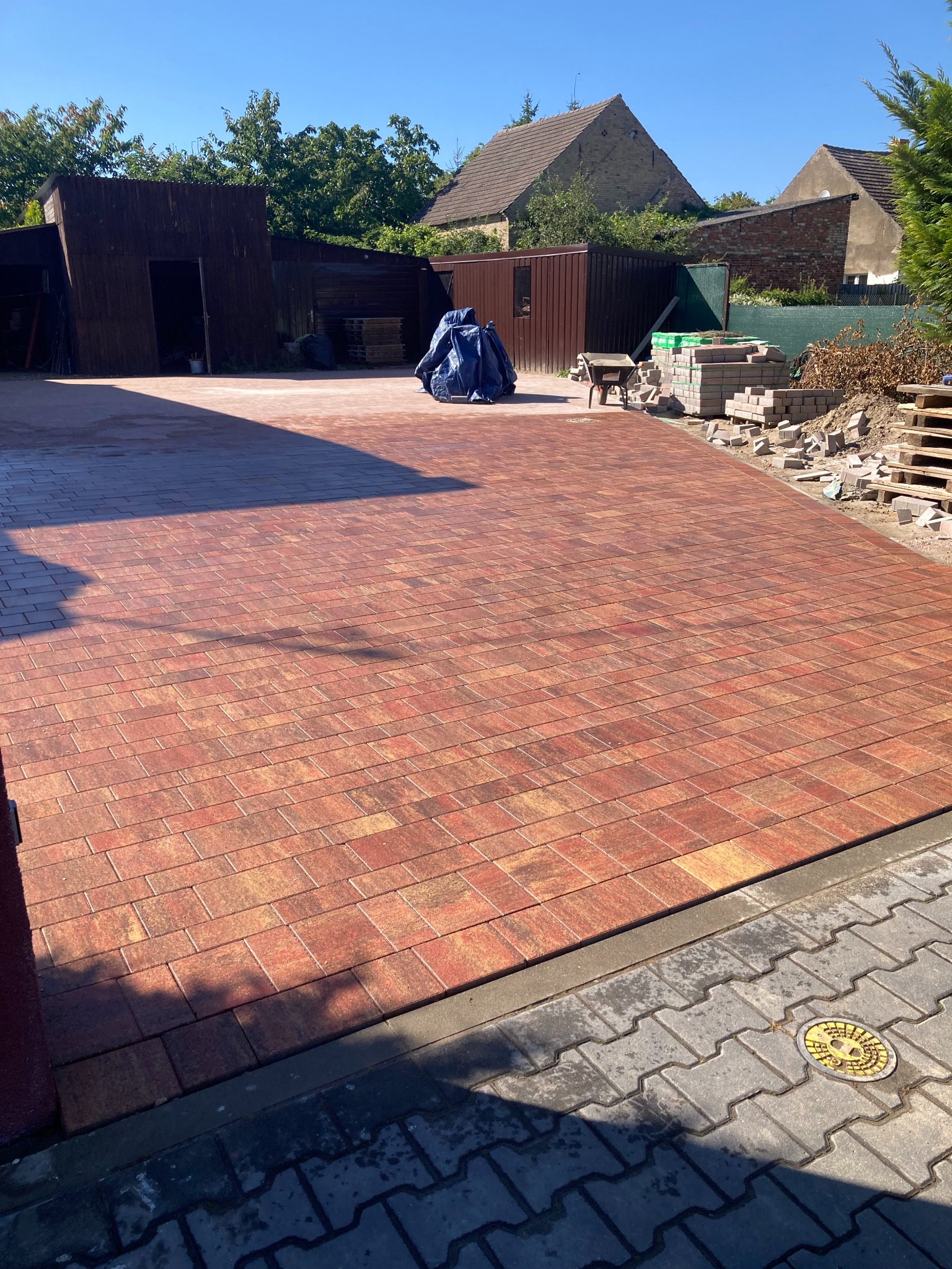 Ukończony podjazd z czerwonej kostki brukowej w jodełkę, widoczne palety z kostką, budynki gospodarcze w tle i studzienka kanalizacyjna na dole zdjęcia.