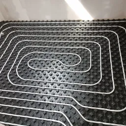 Białe rury ogrzewania podłogowego ułożone spiralnie na czarnej macie izolacyjnej w surowym wnętrzu, widoczne fragmenty instalacji przy ścianie.