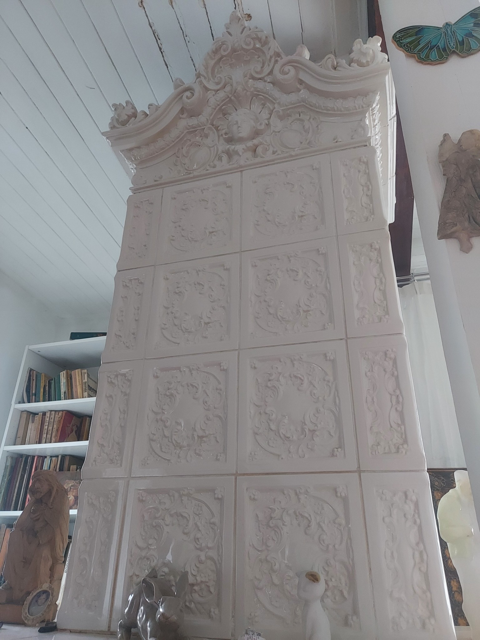 Wysoki, biały piec kaflowy z dekoracyjnymi płaskorzeźbami, widziany z dołu, z elementami wystroju wnętrza widocznymi w tle, w tym regał z książkami, drewniana rzeźba i dekoracja motyla na ścianie.
