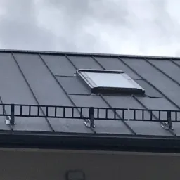 Szary dach z blachy trapezowej z zamontowanym oknem dachowym i barierką śniegową w kształcie drabinki, widoczne elementy komina na tle pochmurnego nieba.