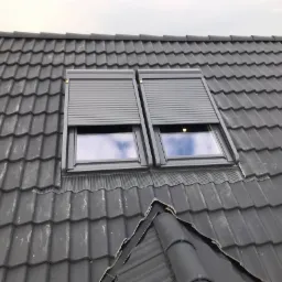 Dwa okna dachowe z szarymi roletami zewnętrznymi na ciemnoszarym dachu z dachówki ceramicznej, widoczne odbicie nieba w szybach.
