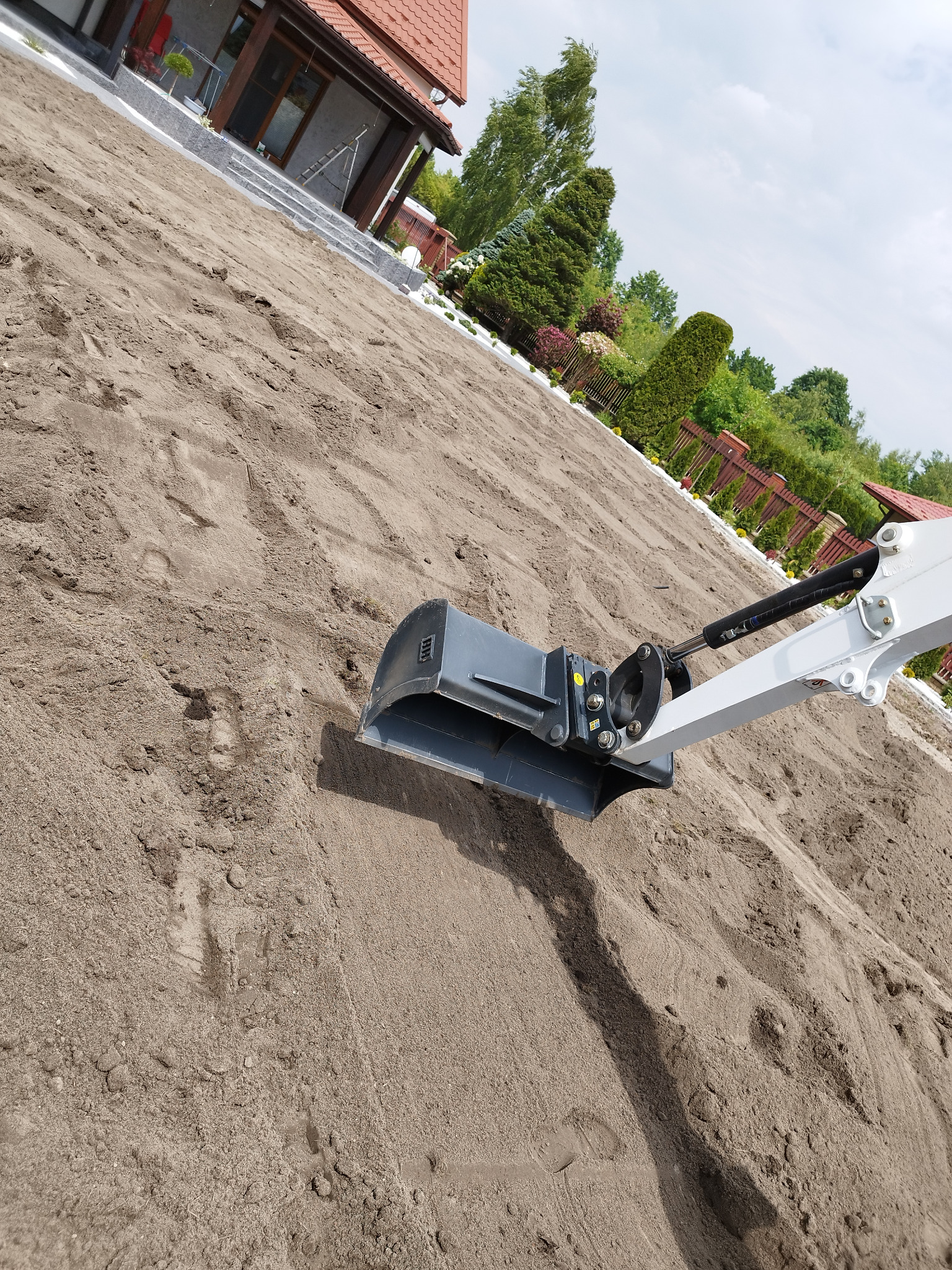 Mini koparka z łyżką wyrównuje teren piaskiem w ogrodzie przy domu jednorodzinnym, widoczne ślady gąsienic na piasku.