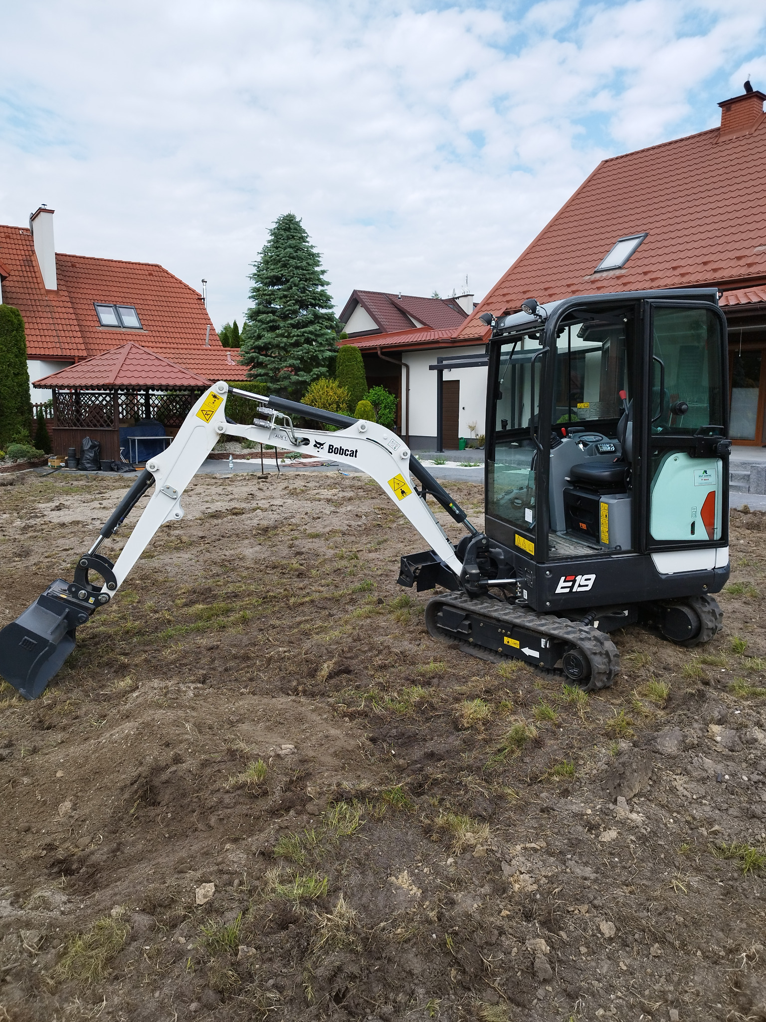 Kompaktowa minikoparka Bobcat E19 na gąsienicach, stojąca na nierównym, częściowo wykopanym terenie z widocznymi śladami pracy, w tle domy jednorodzinne z czerwonymi dachami i zielenią.