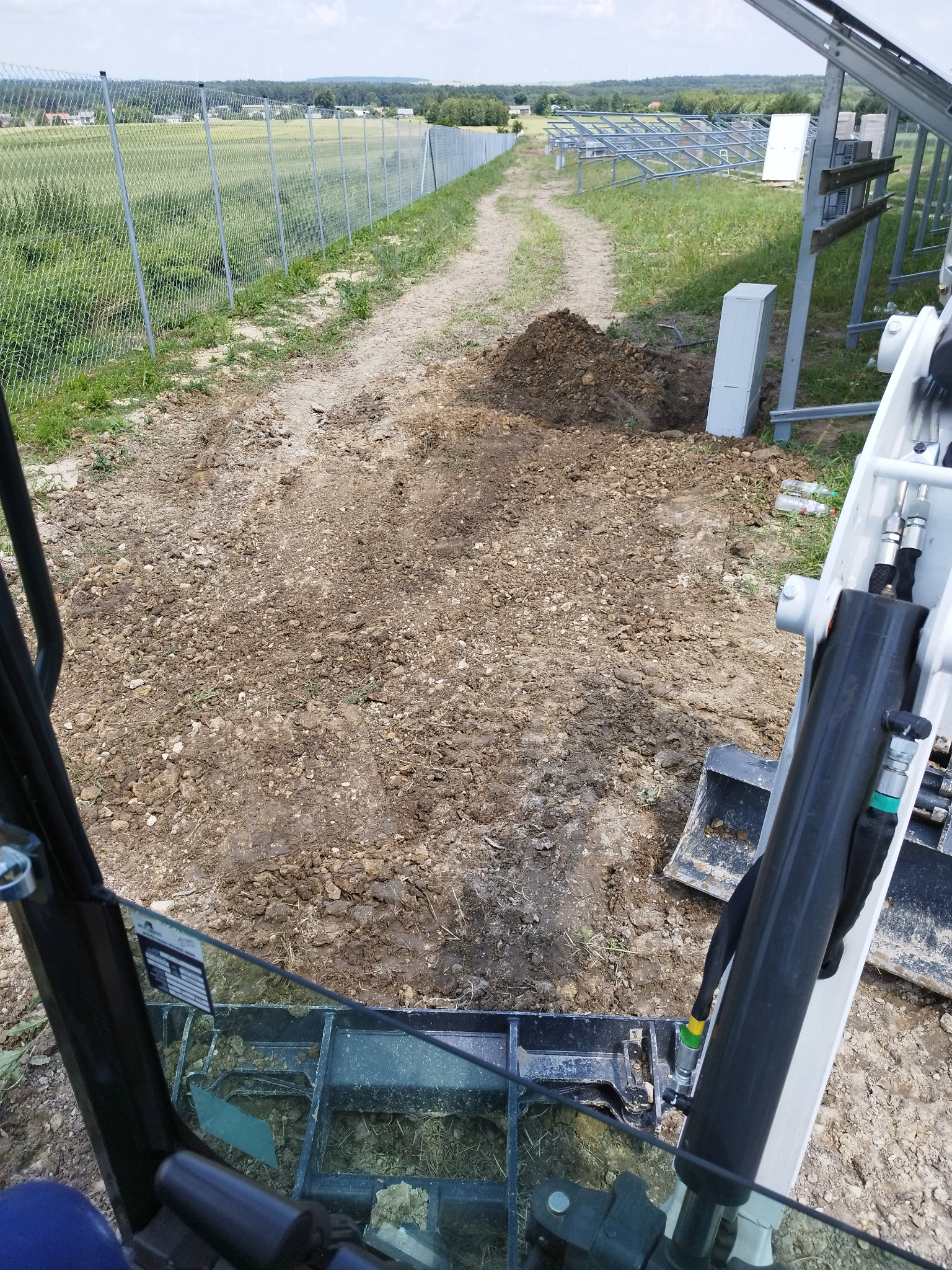 Widok z kabiny minikoparki na wykop ziemi pod instalację, z widocznymi elementami konstrukcji pod panele słoneczne w tle, obok polnej drogi i ogrodzenia z siatki.