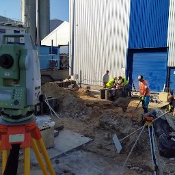 ŁUKASZ MARSZAŁEK USŁUGI GEODEZYJNO-PROJEKTOWE - Praca na budowie: teodolit na statywie, w tle pracownicy przy wykopie fundamentowym i hala z niebieskimi akcentami. Słoneczny dzień.
