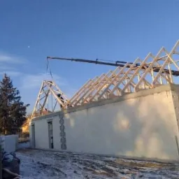 Montaż drewnianej konstrukcji dachu na budynku z bloczków betonowych, widoczny dźwig, zimowa sceneria z resztkami śniegu.