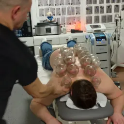 Pacjent leży na brzuchu na kozetce z przyczepionymi bańkami próżniowymi na plecach, terapeuta masuje jego ramię, w tle widoczne plakaty z punktami spustowymi, lampa solna i sprzęt medyczny.