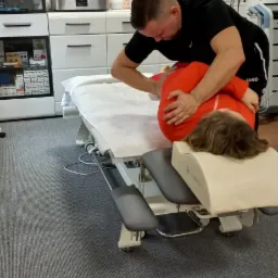 Fizjoterapeuta wykonuje manualną terapię kręgosłupa lędźwiowego u pacjentki leżącej na profesjonalnym stole rehabilitacyjnym w gabinecie z planszami anatomicznymi.