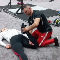 Mężczyzna w sportowym stroju rozciąga nogę leżącego na podłodze sportowca na tle siłowni z innymi osobami i sprzętem do ćwiczeń.