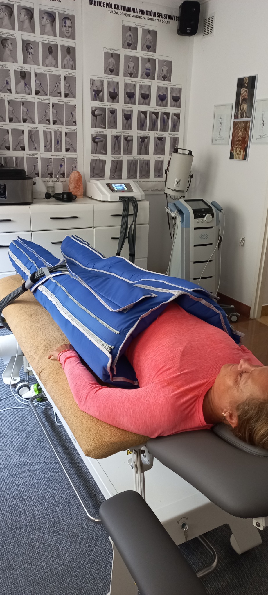 Kobieta w różowej koszulce leży na stole do zabiegów, poddana presoterapii z wykorzystaniem specjalnego kombinezonu pneumatycznego na nogach i tułowiu; w tle widoczne tablice anatomiczne...