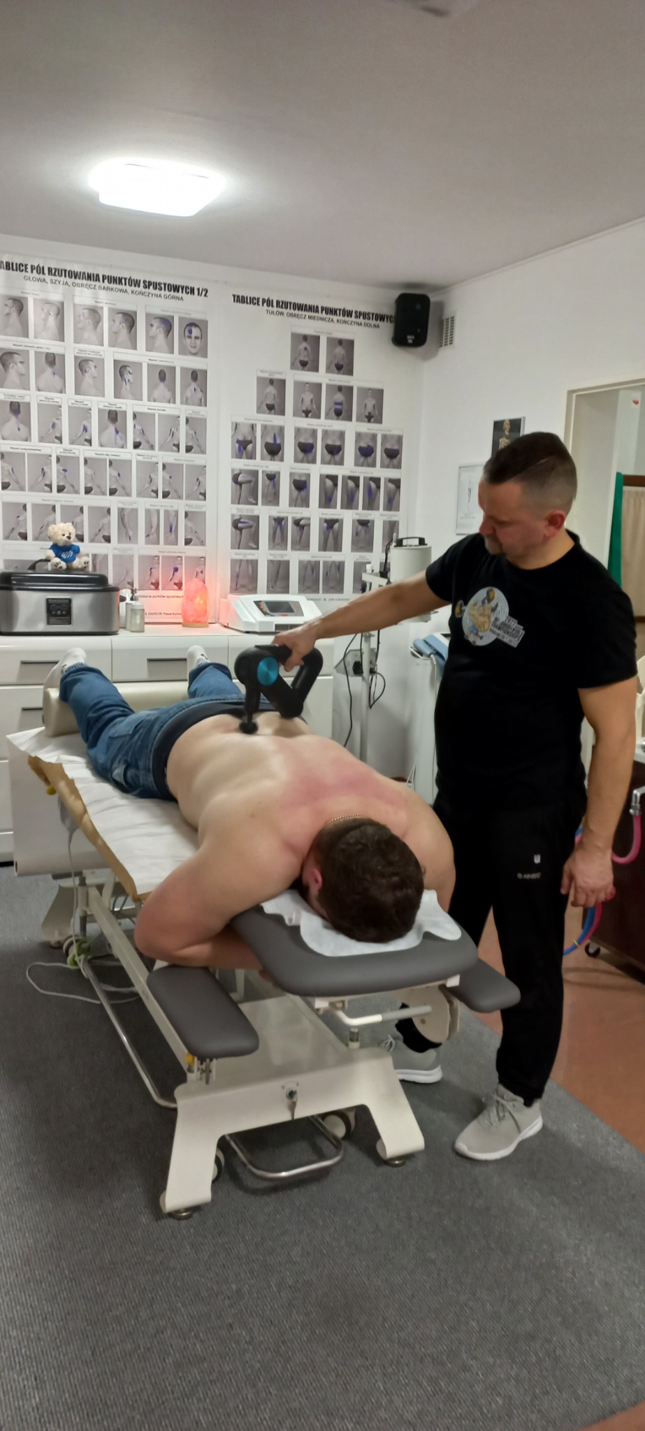 Pacjent leżący na kozetce podczas masażu pleców urządzeniem wibracyjnym; w tle widoczne tablice z punktami spustowymi.