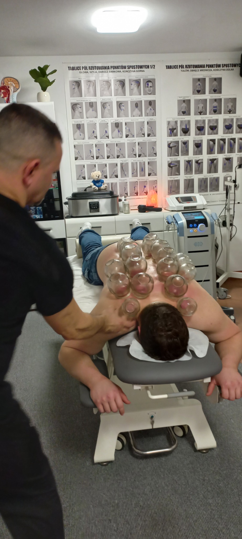 Pacjent leży na brzuchu na kozetce z przyczepionymi bańkami próżniowymi na plecach, terapeuta masuje jego ramię, w tle widoczne plakaty z punktami spustowymi, lampa solna i sprzęt medyczny.