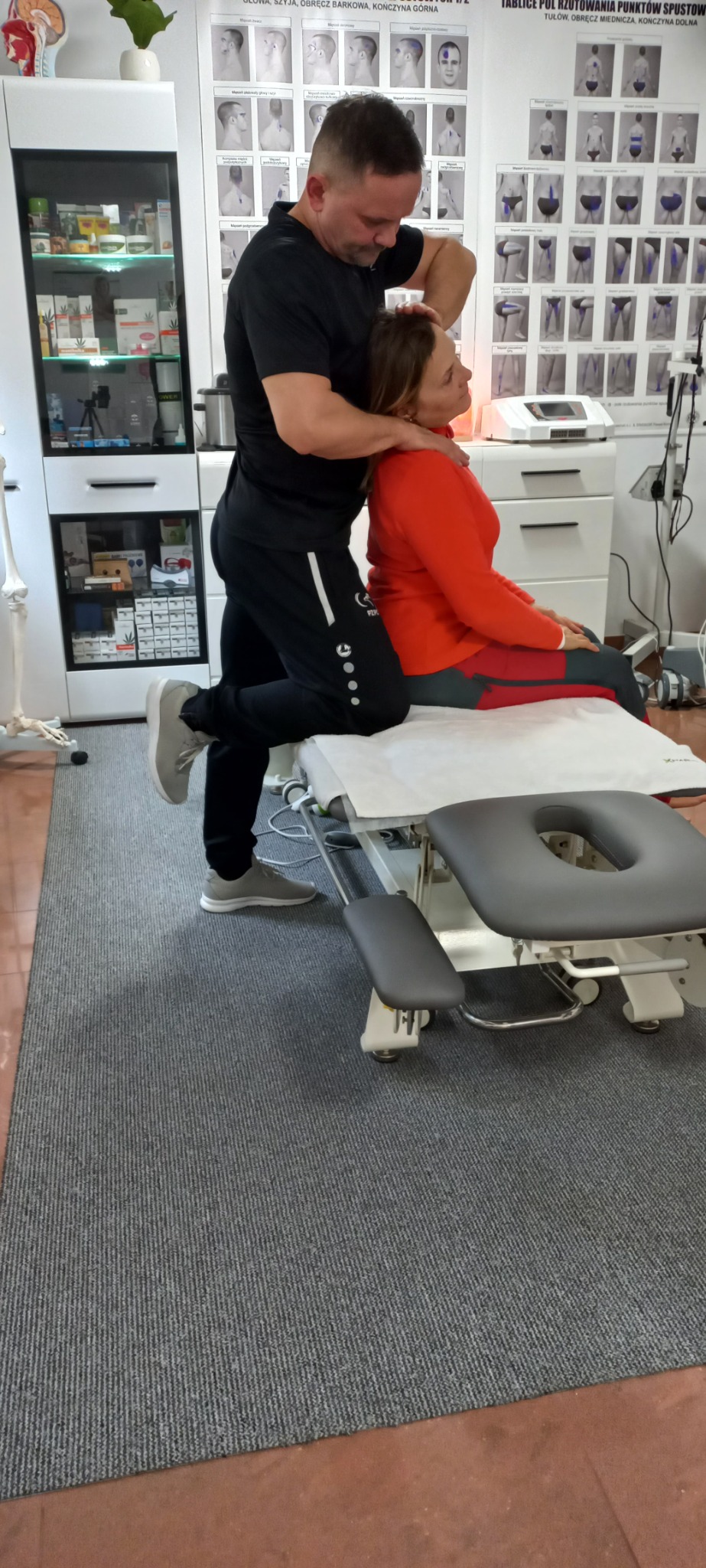 Specjalista w czarnym stroju wykonuje terapię manualną szyi pacjentki siedzącej na profesjonalnym stole do masażu; widoczne plakaty anatomiczne na ścianie i szafka z produktami w tle.