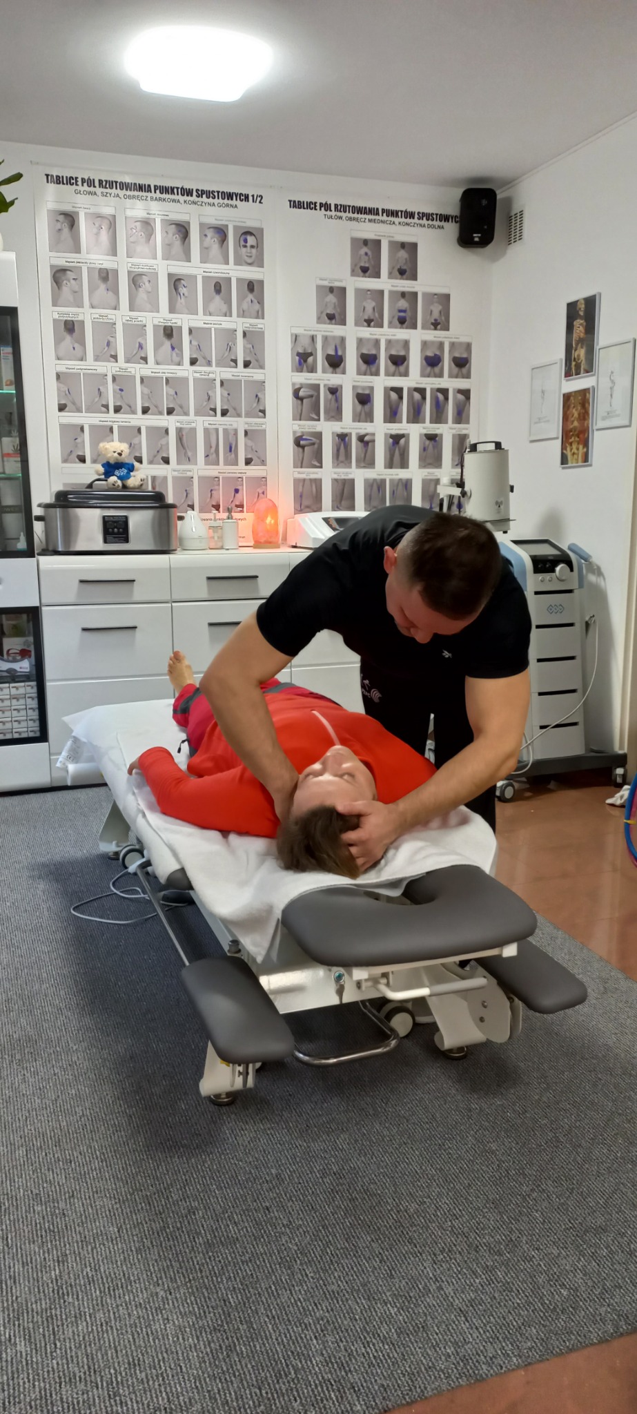 Pacjentka w czerwonej bluzie leży na specjalistycznym łóżku do rehabilitacji, terapeuta w czarnym ubraniu wykonuje manualny zabieg na szyi i głowie, w tle widoczne plakaty z mapami punktów...