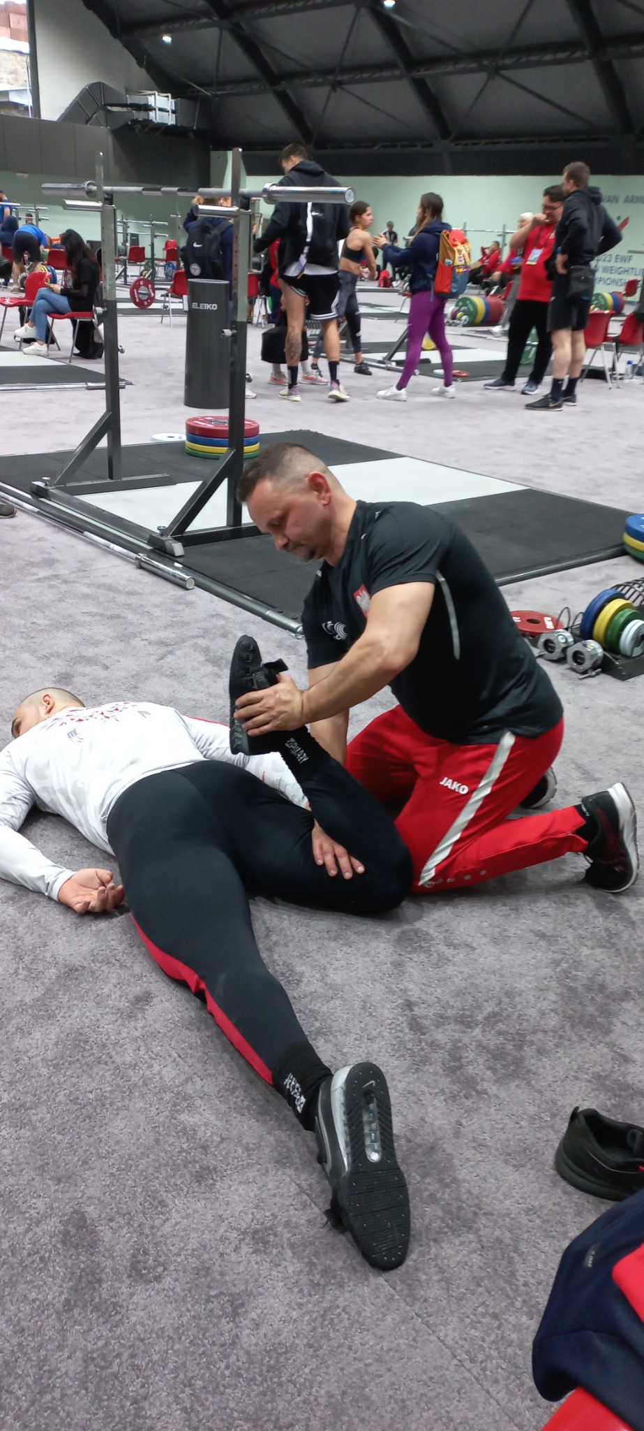 Mężczyzna w sportowym stroju rozciąga nogę leżącego na podłodze sportowca na tle siłowni z innymi osobami i sprzętem do ćwiczeń.