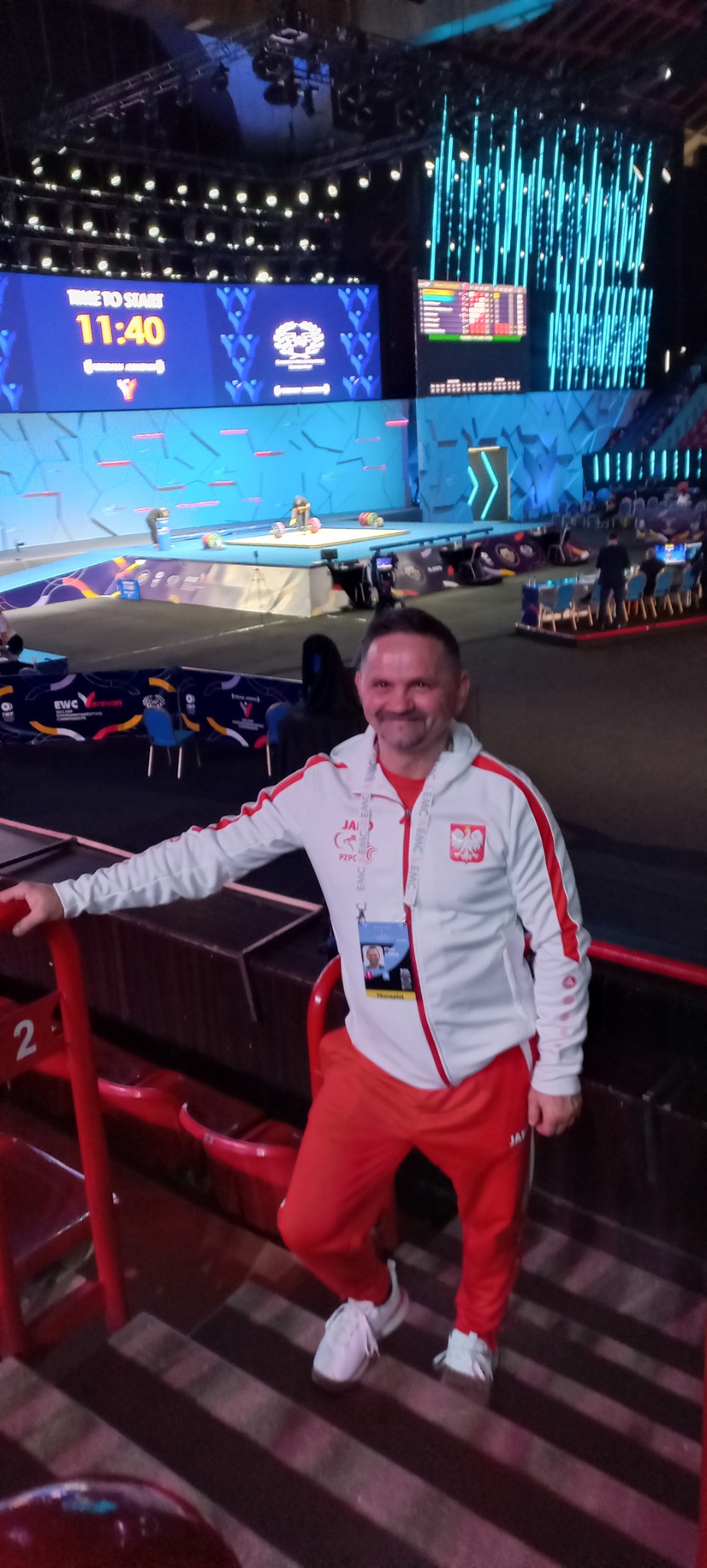 Mężczyzna w dresie reprezentacji Polski na zawodach podnoszenia ciężarów, na widowni, w tle scena z zawodnikami i tablica wyników.