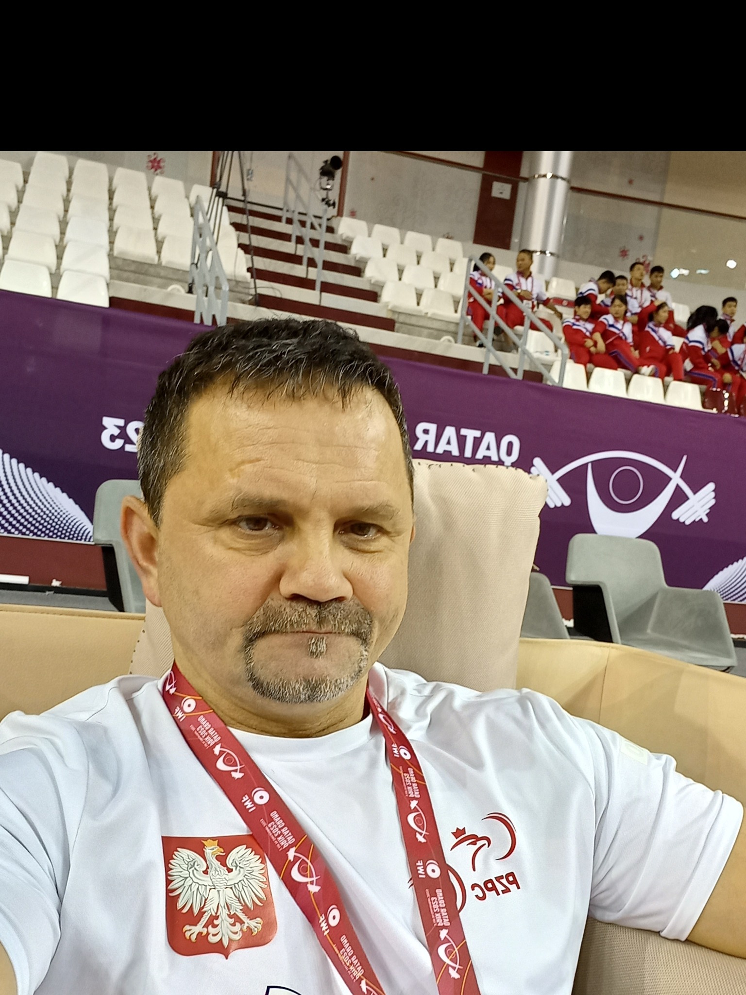 Portret mężczyzny w koszulce z godłem Polski i logiem PFRON, siedzącego na tle trybun sportowych podczas wydarzenia w Katarze.
