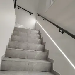 Wewnętrzne schody wyłożone szarymi płytkami z czarną, minimalistyczną poręczą zamocowaną do ściany, oświetlone listwą LED wzdłuż ściany.