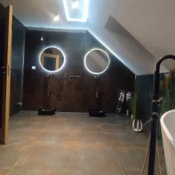 Nowoczesna łazienka na poddaszu z dwoma okrągłymi lustrami z podświetleniem LED, ciemnymi płytkami na ścianach i podłodze, czarnymi umywalkami nablatowymi oraz oświetleniem sufitowym w kształcie...