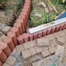 Budowa ścieżki ogrodowej z użyciem okrągłych palisad i kwadratowej kostki brukowej, widoczne narzędzia: miotła, poziomica, ołówek, metrówka, nóż, fragment buta pracownika.