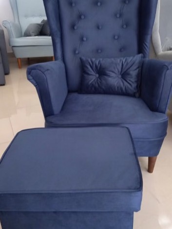 Granatowy fotel uszak z podnóżkiem obity miękkim welurem, z dekoracyjnym pikowaniem na oparciu i poduszce, na tle jasnej podłogi i innych mebli.