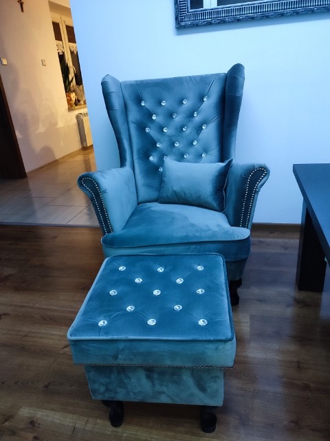 Turkusowy fotel typu uszak z podnóżkiem, obity welurem i zdobiony kryształkami, ustawiony na parkiecie w jasnym wnętrzu.