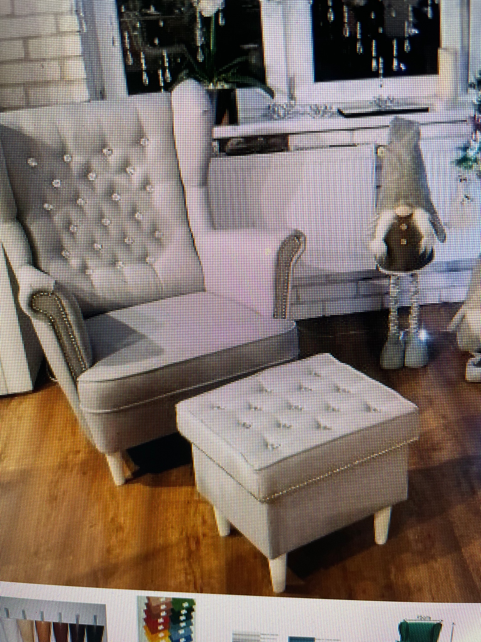 Elegancki, szary fotel typu uszak z pikowanym oparciem i podnóżkiem, wykończony srebrnymi detalami, na tle jasnego wnętrza z dekoracjami.