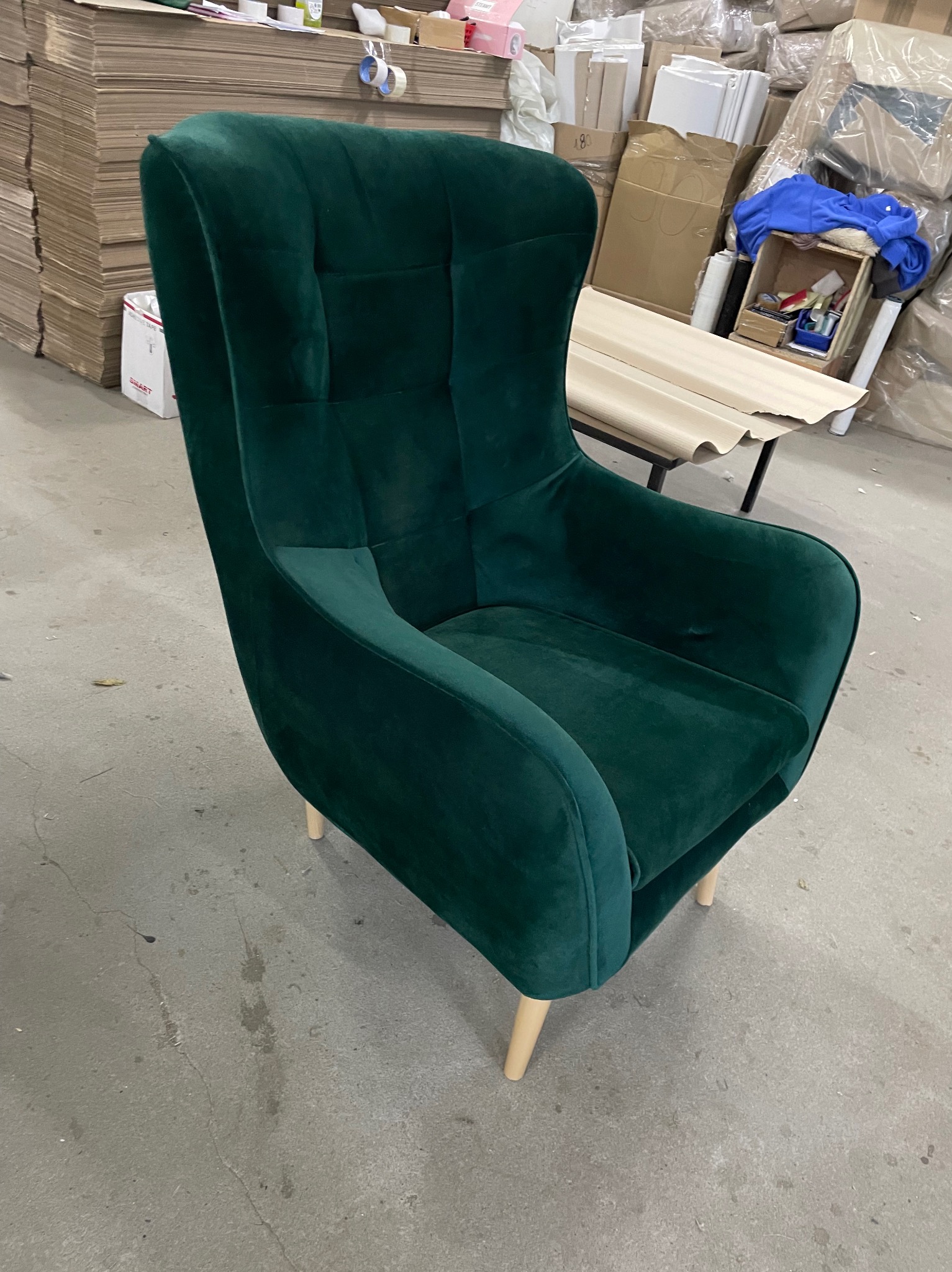 Wysokie, tapicerowane, zielone krzesło z guzikami na oparciu, na jasnych, drewnianych nóżkach, sfotografowane w magazynie lub warsztacie.