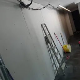 Wnętrze w trakcie prac wykończeniowych: ściany z płyt gipsowo-kartonowych, aluminiowa drabina przystawiona do ściany, wiadro z zaprawą, widoczne kable pod sufitem.