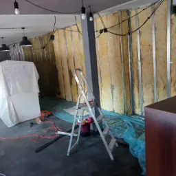 Ściana w trakcie izolacji wełną mineralną, widoczne metalowe profile, kable elektryczne, drabina aluminiowa, podłoga zabezpieczona folią, elementy wyposażenia wnętrza przykryte folią malarską.