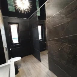 Nowoczesna łazienka z czarnymi płytkami, drewnopodobną podłogą i dekoracyjną lampą sufitową w kształcie gwiazdy.