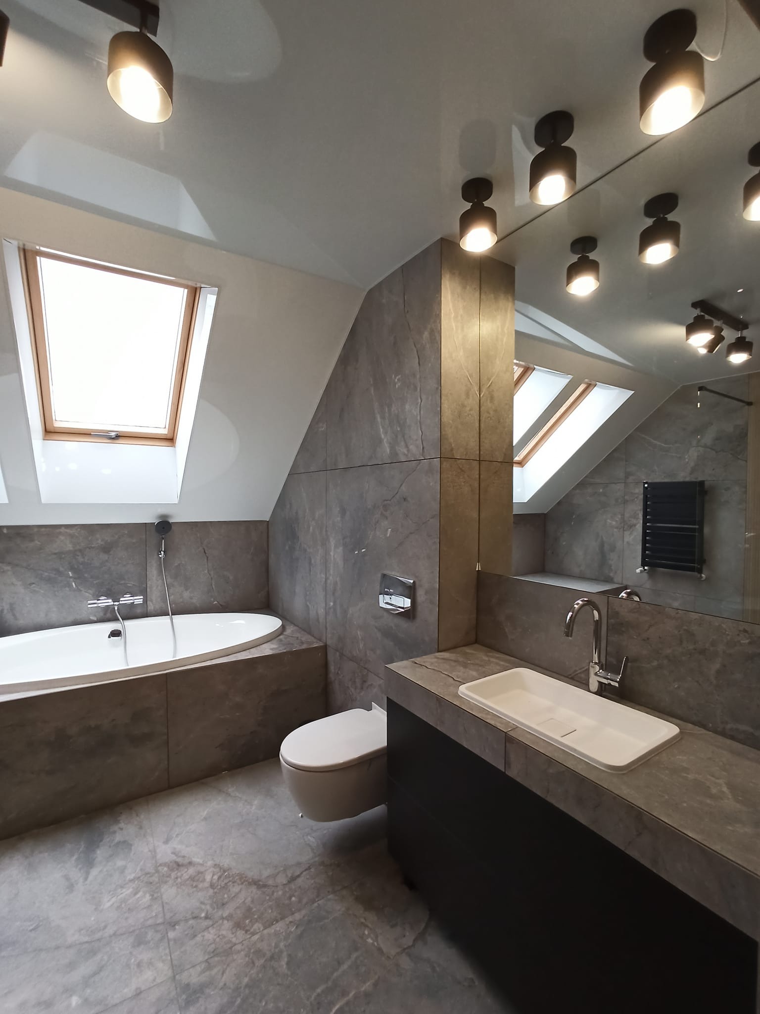 Nowoczesna łazienka na poddaszu z oknem dachowym, szarą płytką imitującą kamień na ścianach i podłodze, białą wanną i umywalką oraz czarnymi szafkami pod umywalką, oświetlona reflektorami.