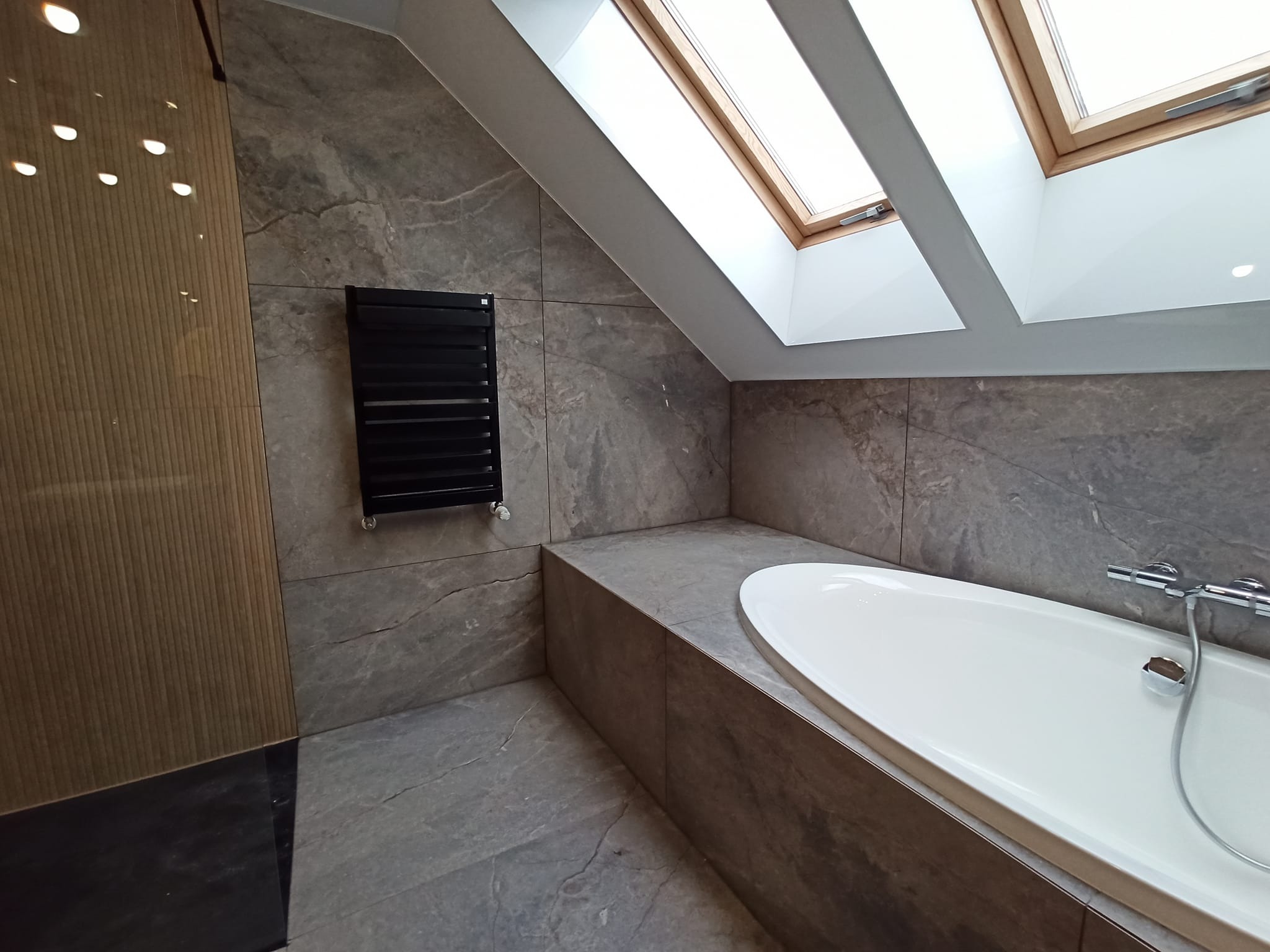 Nowoczesna łazienka na poddaszu z szarymi płytkami imitującymi kamień, białą wanną, czarnym grzejnikiem drabinkowym i oknami dachowymi.