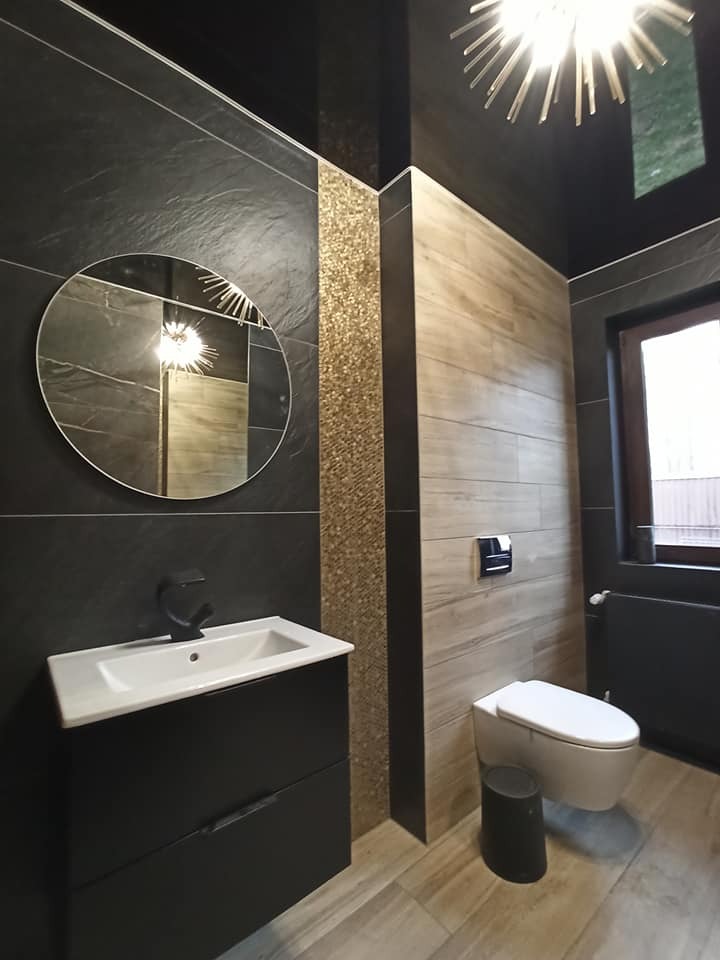 Nowoczesna łazienka z czarnymi płytkami imitującymi kamień, drewnopodobnymi panelami, okrągłym lustrem i złotą mozaiką akcentującą narożnik. Czarna szafka pod umywalką i wisząca toaleta.