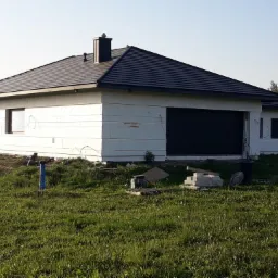 Niedokończony dom jednorodzinny z ociepleniem ścian zewnętrznych w postaci białych płyt styropianowych, czarnym dachem z dachówki ceramicznej i fragmentem podniesionego terenu wokół budynku.