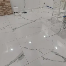 Błyszcząca, biała podłoga z płytek imitujących marmur, ułożona w salonie z ceglaną ścianą, widoczny robot sprzątający i białe krzesła.