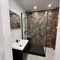 Nowoczesna łazienka z kabiną prysznicową typu walk-in z czarnymi ramami, umywalką nablatową na czarnej szafce z złotymi uchwytami i płytkami imitującymi marmur na ścianach.