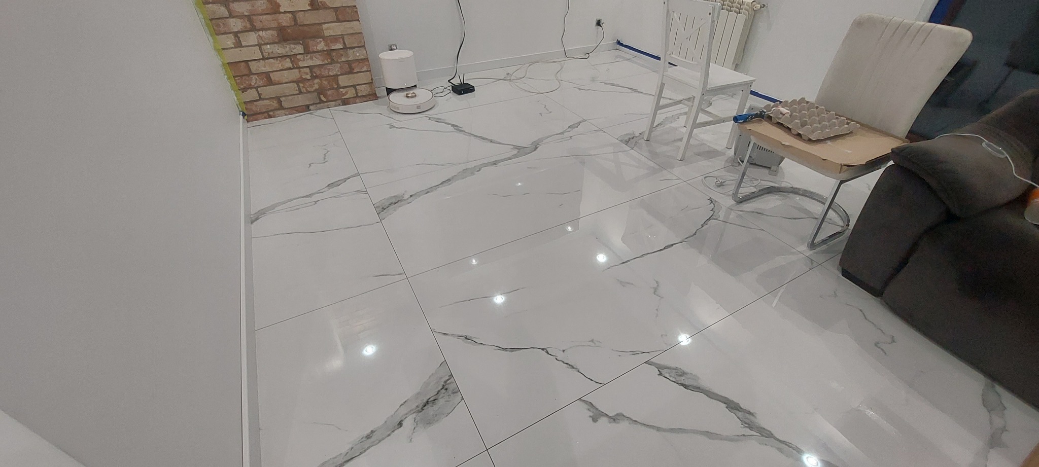 Błyszcząca, biała podłoga z płytek imitujących marmur, ułożona w salonie z ceglaną ścianą, widoczny robot sprzątający i białe krzesła.