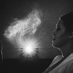 Czarno-białe zdjęcie profilu kobiety, na którą rozpylana jest mgiełka kosmetyczna; widoczna ręka trzymająca opakowanie produktu, oświetlone od tyłu delikatnym światłem.