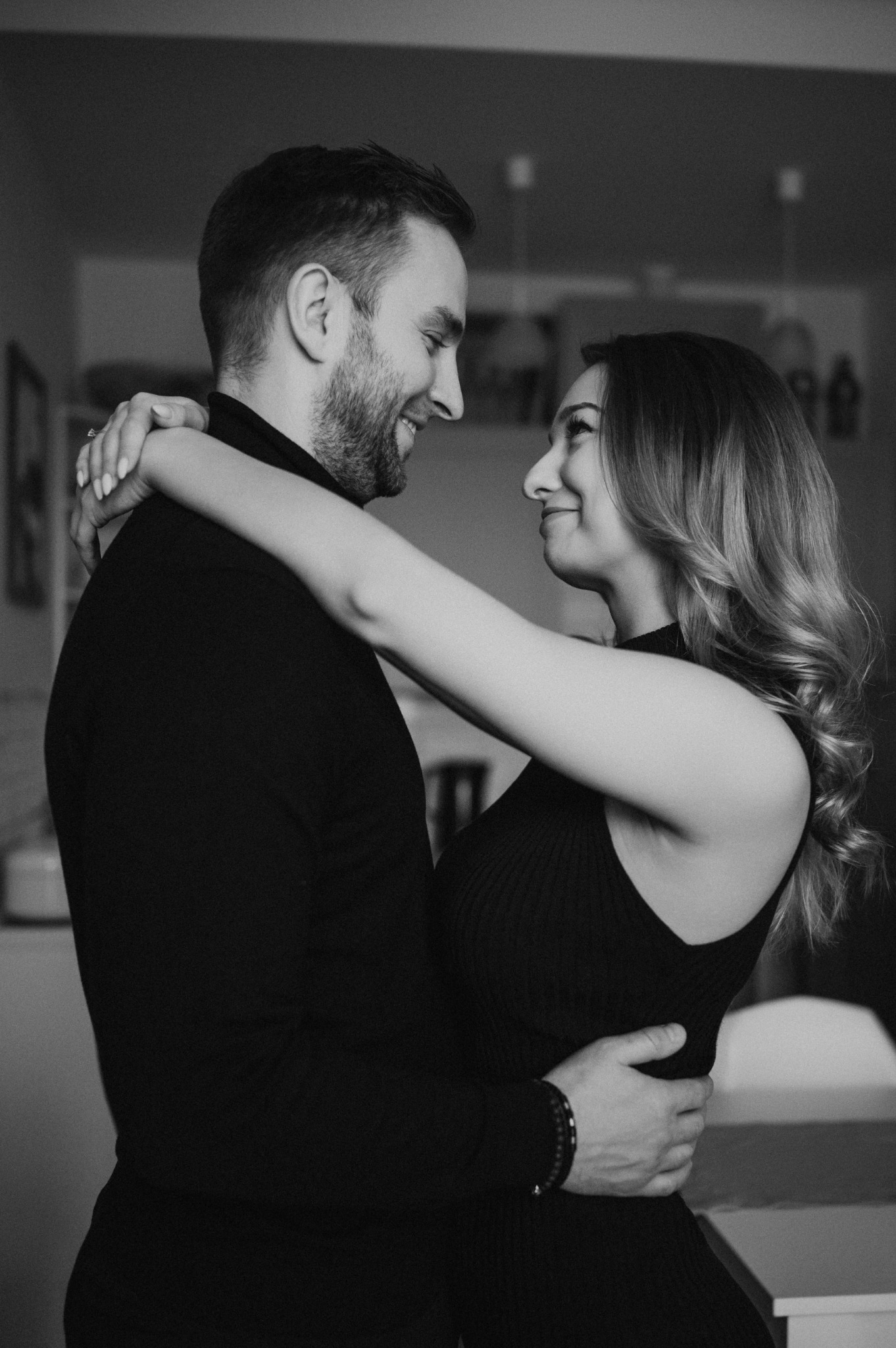 Czarno-białe zdjęcie uśmiechniętej pary w objęciach, kobieta z falowanymi włosami i pierścionkiem na palcu, mężczyzna z brodą, oboje w ciemnych ubraniach.